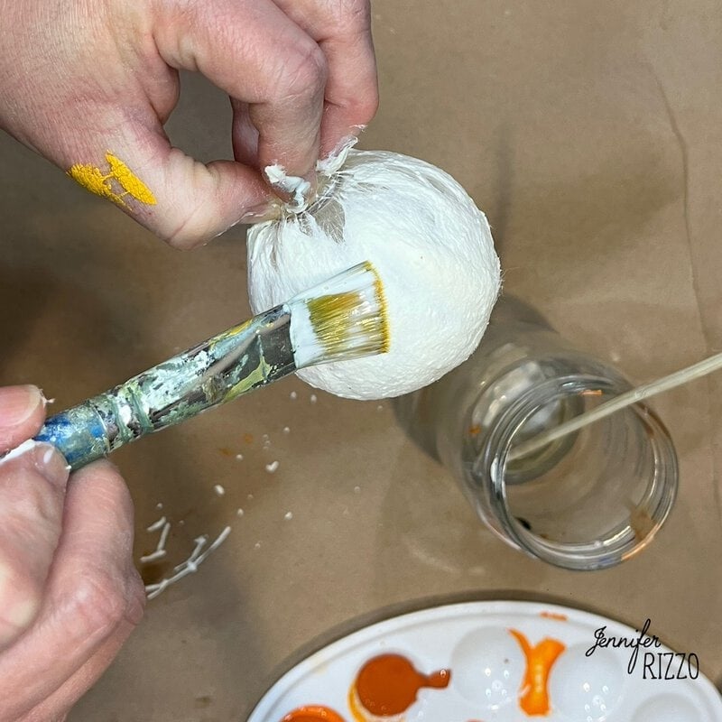 Person painting a white ornament with a brush, holding it above a jar. Painting supplies, including orange paint and a palette, are visible.