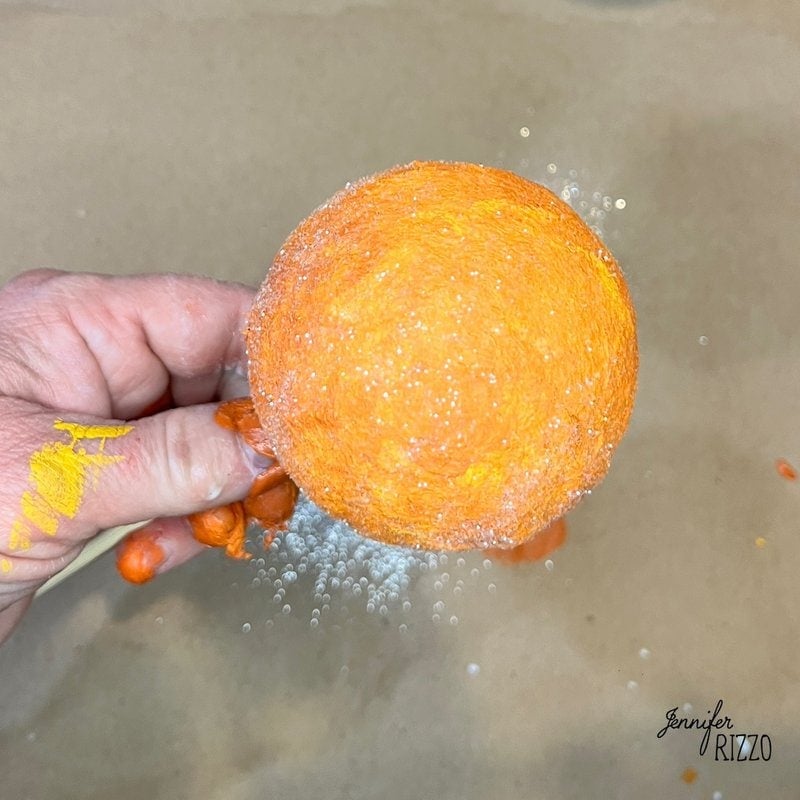 Hand holds an orange foam ball covered in glitter, with some paint on fingers. Brown surface in background.