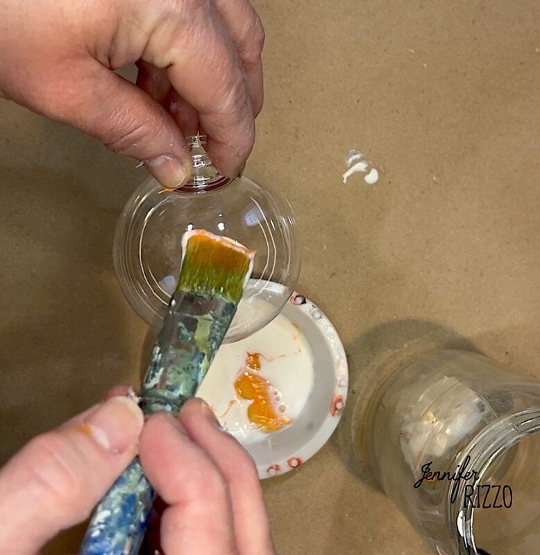 Hands holding a clear ornament and painting with a brush over a small dish with paint.