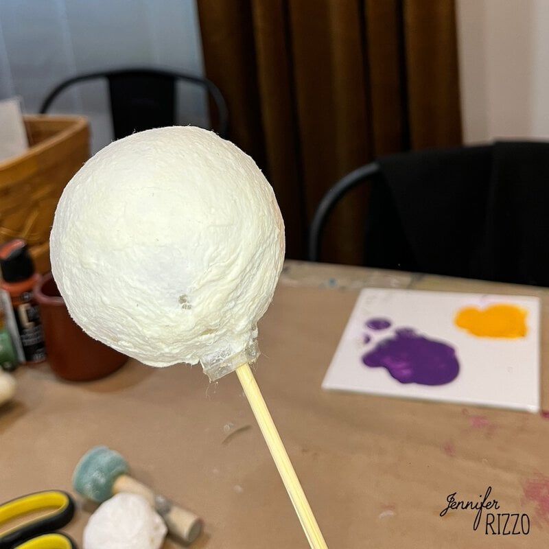 A white ornament covered in spun cotton batting anf glue on a stick is being held over a table with purple and orange paint on a palette in the background.