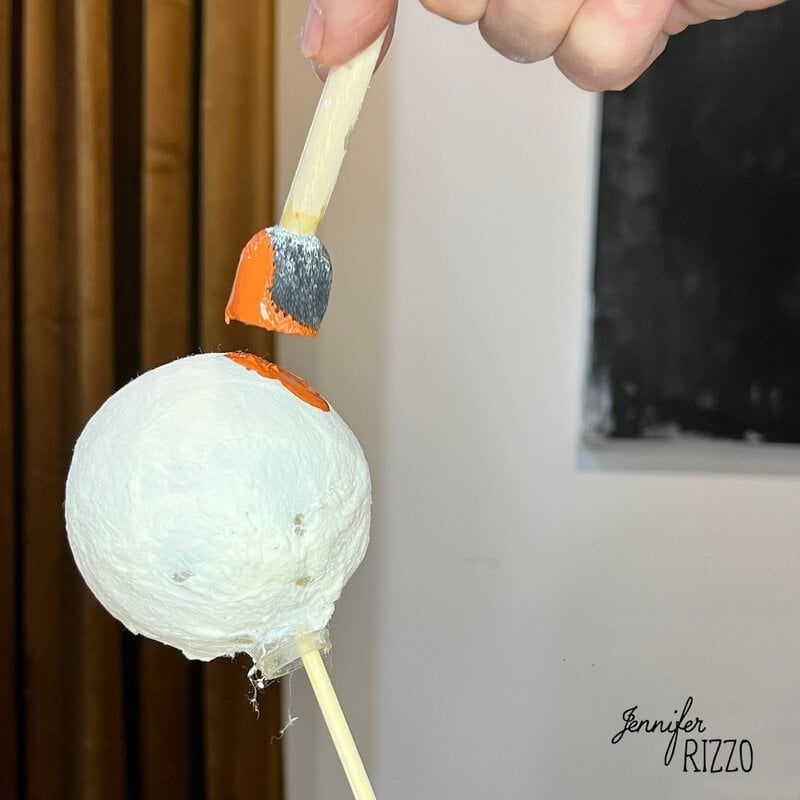 A hand paints an orange circle onto a white foam ball mounted on a stick.