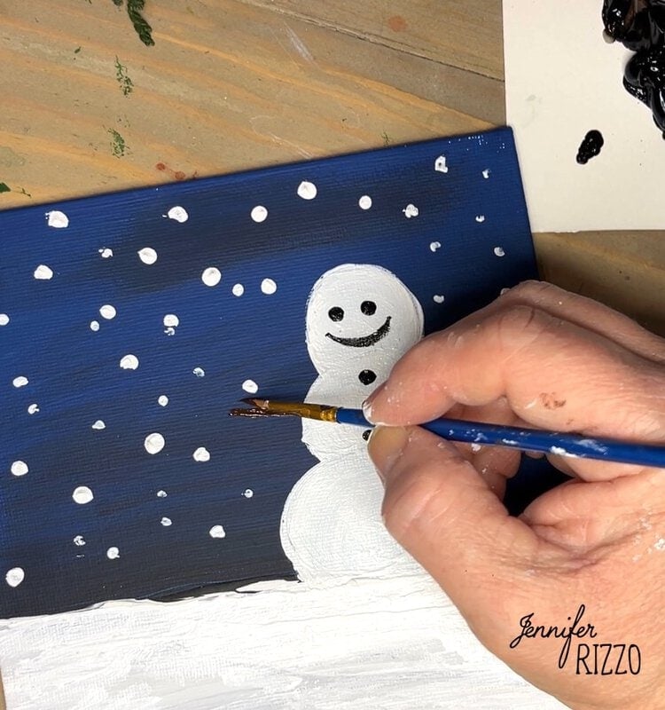 A person paints a smiling snowman on a dark blue canvas with falling snowflakes, using a paintbrush and black and white paint.