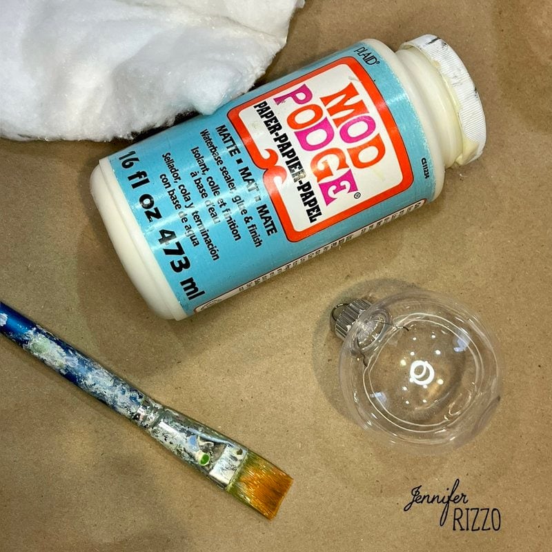 A jar of Mod Podge, a paintbrush, a clear ornament, and cotton on a brown surface.