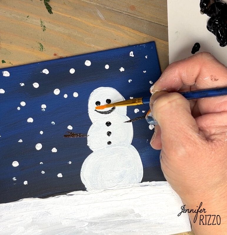 A person paints the carrot nose of a snowman on a winter-themed canvas with a brush.