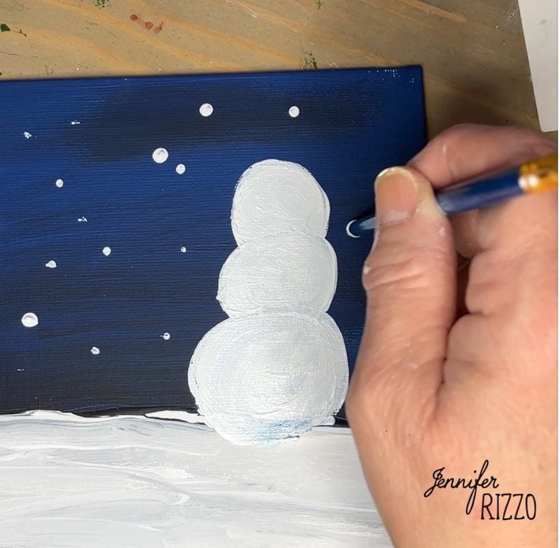 A hand paints a white snowman and snowflakes on a dark blue background. Artist signature "Jennifer Rizzo" is in the bottom corner.