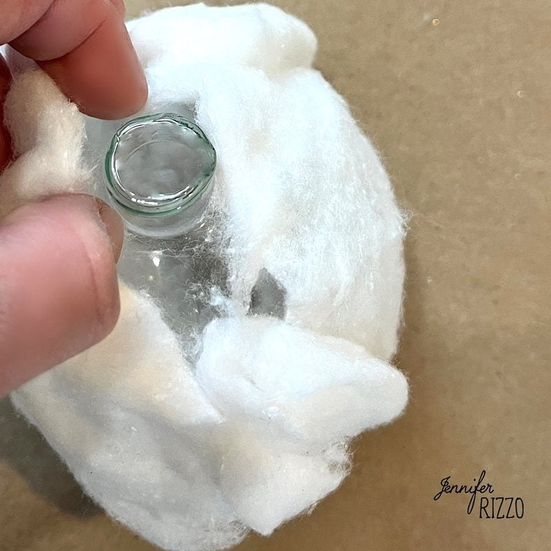 Hand holding a small glass bottle nestled in white cotton, with a beige background and a watermark in the corner.
