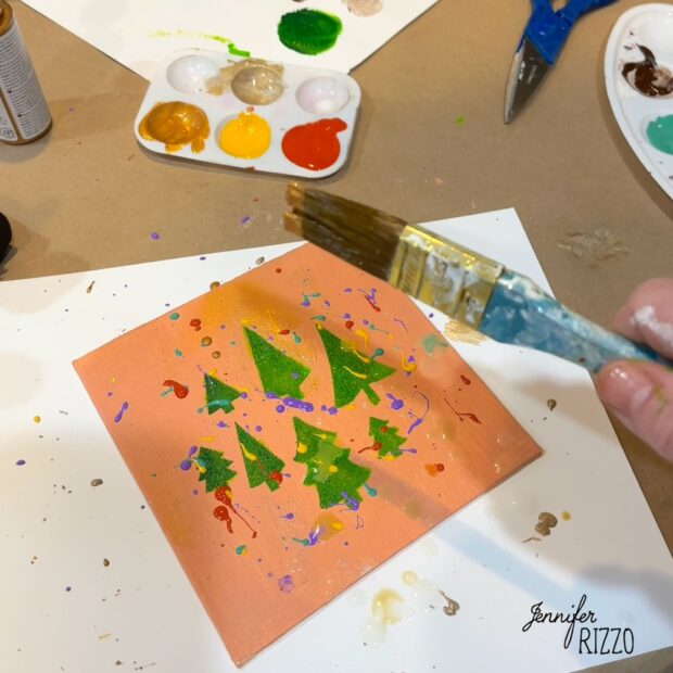 A person holds a paintbrush over a square canvas with painted green Christmas trees and colorful splatters. Paint supplies and scissors are scattered around on a table.