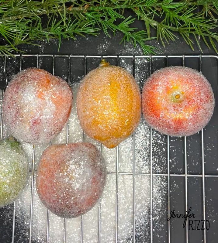 Frosted apples and a lemon on a cooling rack, dusted with powdered sugar; evergreen branches in the background.