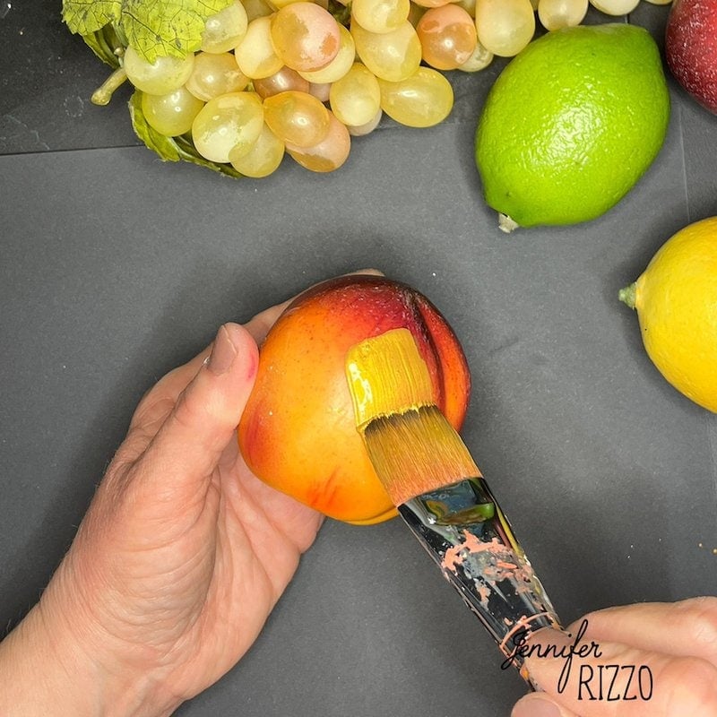 A hand paints an apple with a brush. Grapes and citrus fruits are scattered around on a black surface.