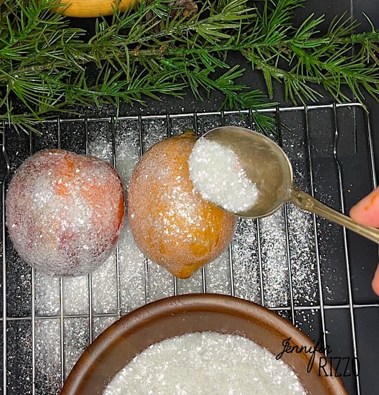 Two oranges on a rack are being sprinkled with white sugar from a spoon. Evergreen sprigs are in the background to Make faux sugared fruits