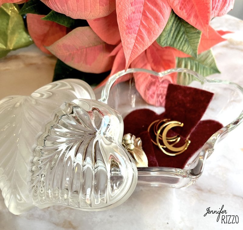 A glass heart-shaped dish with gold earrings and a ring on red felt, next to a pink and green poinsettia plant.