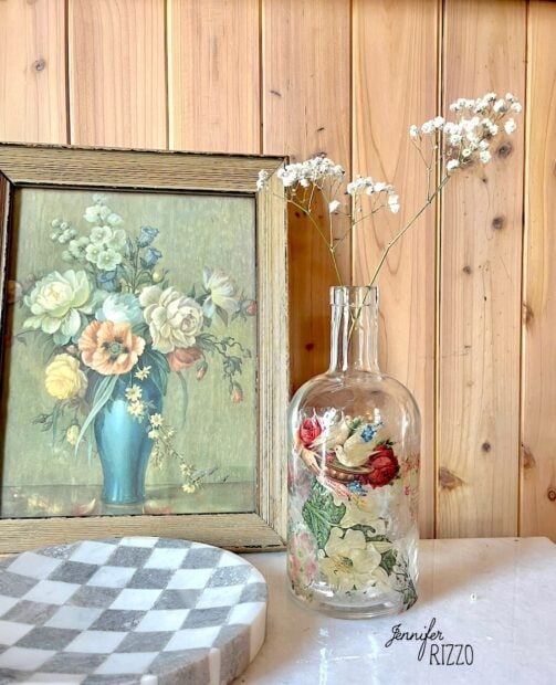A decorated glass bottle with dried flowers sits next to a framed floral painting on a wooden surface, while DIY stickers crafted with tape lend charm to a nearby checkered pattern round object.
