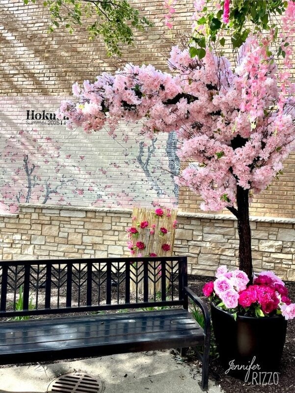A black bench sits in front of a brick wall mural with pink flowers and a faux pink flowering tree, next to a pot of vibrant pink and white flowers, offering a peaceful Hokusai in Lisle-inspired scene.