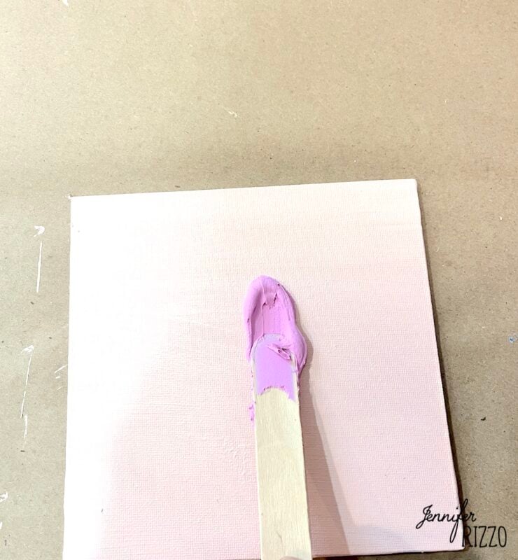 A wooden stick spreads light pink paint on a small square canvas, placed on a brown work surface.