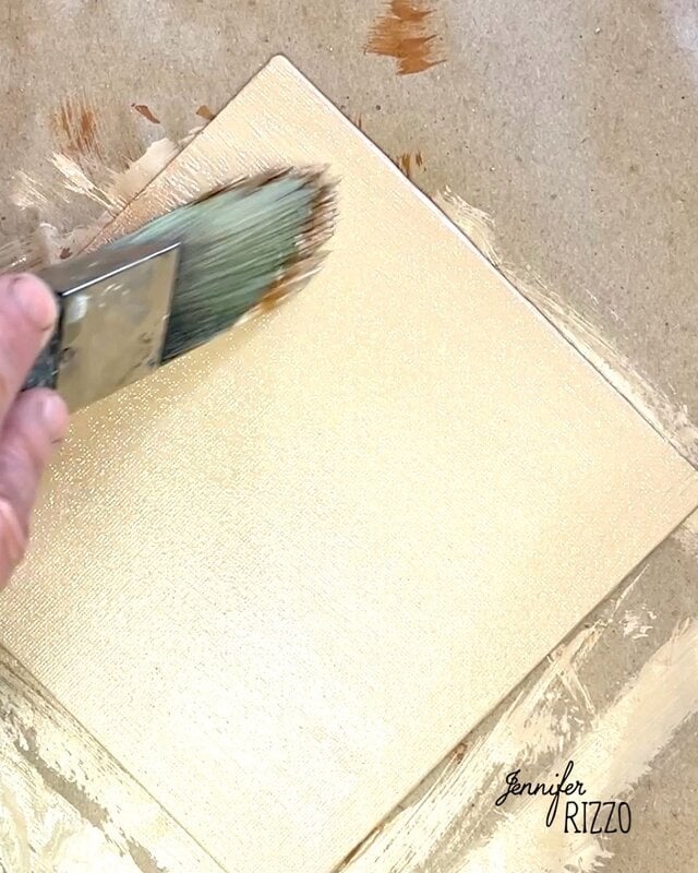 A hand uses a paintbrush to apply a light, shimmery paint onto a square canvas placed on a brown surface.