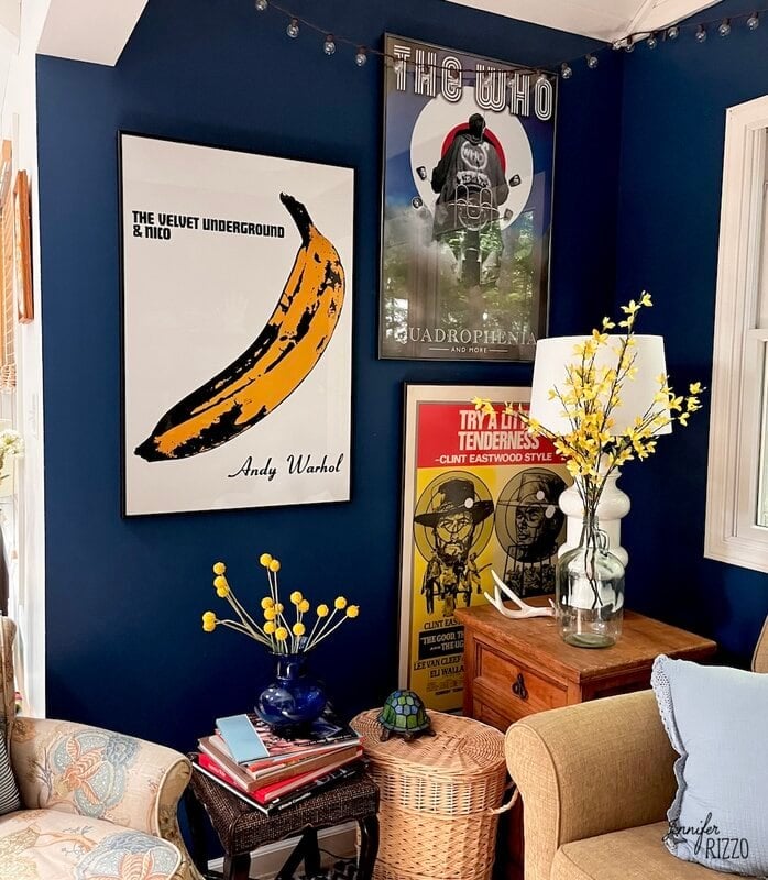 A cozy corner with dark blue walls features three framed posters, a small table with a yellow flower arrangement, books, and a wicker basket.