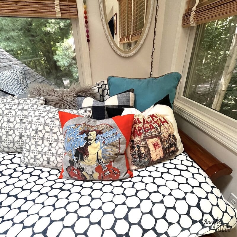 A bed with patterned sheets, decorative pillows including one with an Elvis Presley design, is positioned next to windows with a forest view and a round wall mirror above.