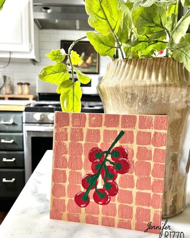A small painting of red tomatoes on a vine is displayed on a kitchen counter next to a potted plant, with a stove and white tile backsplash in the background.