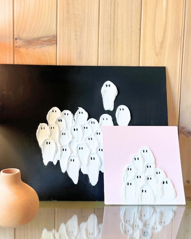 Two paintings of white cartoon ghosts with black eyes are displayed against a wooden wall; one has a black background, the other a pink background. A small beige vase sits in the foreground.