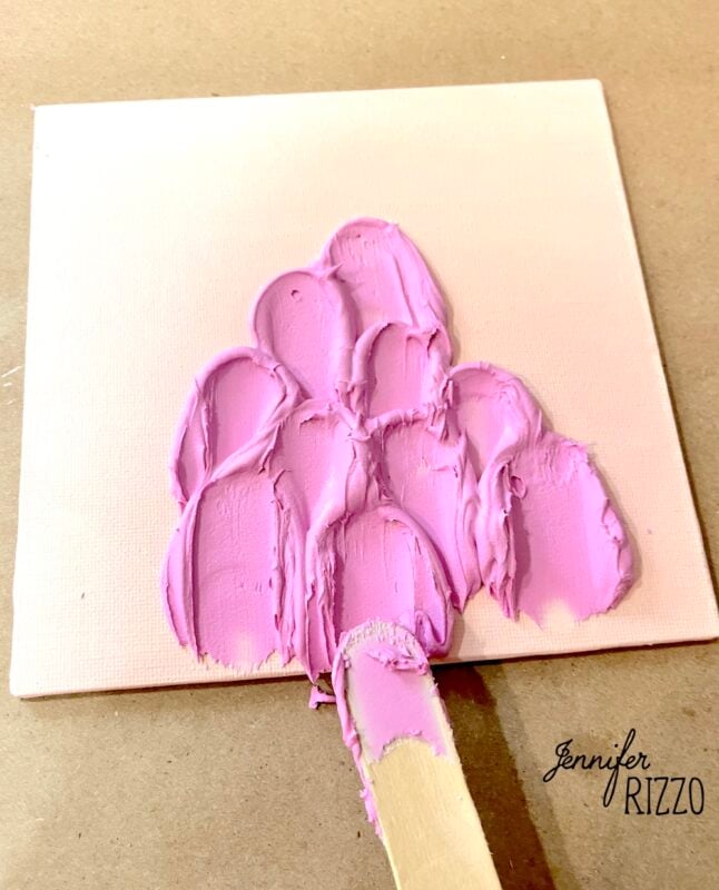 A palette knife spreads thick pink paint in a textured pattern on a small canvas, with the name "Jennifer Rizzo" visible in the corner.