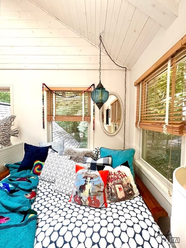 A cozy bed with various decorative pillows and a polka-dot duvet is positioned under windows with wooden blinds, beneath a vaulted white ceiling and a hanging blue lamp.