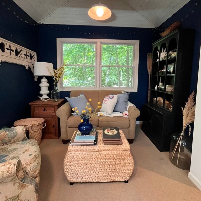 Cozy living room with a beige loveseat, wicker ottoman, side table, and dark bookcase. Window shows greenery outside. Blue walls and neutral decor accents throughout.
