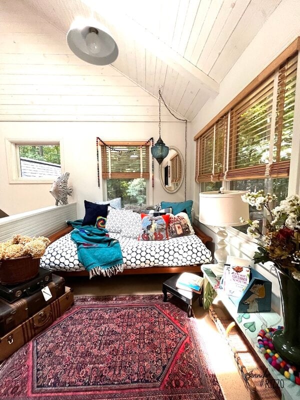 Bright, cozy room with a daybed covered in patterned pillows, surrounded by windows with wooden blinds, a rug, side table with books, and a hanging lamp in a Restyled Eclectic Loft Living Area