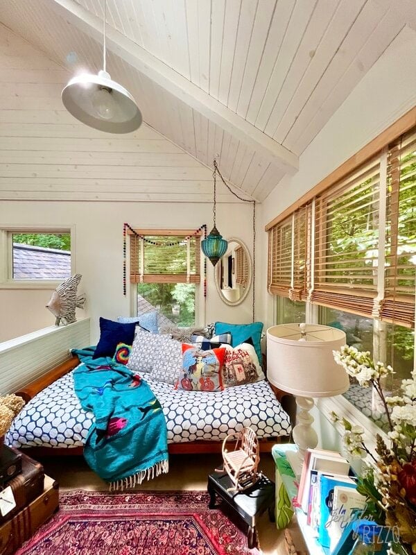 A cozy bedroom with a daybed covered in colorful pillows and a teal throw, a red rug, wooden blinds, and eclectic decor under a white vaulted ceiling.