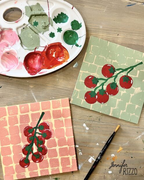 Two square canvases painted with red berry branches sit on a table next to a paint palette with green, pink, and red paints and a paintbrush.