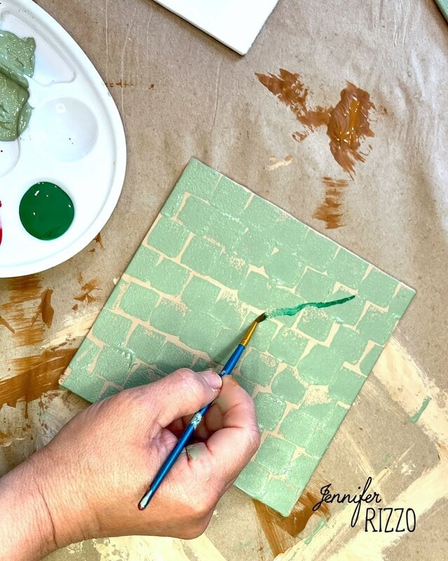 A hand uses a paintbrush to paint green lines on a textured, stencil-like tile. A paint palette with green and white paint is nearby on a paper-covered surface.