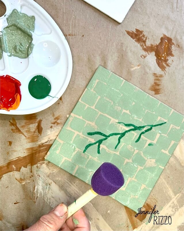 A hand holds a purple foam brush above a painted tile with a green vine on a brick pattern. A paint palette with various colors is nearby on a brown paper surface.