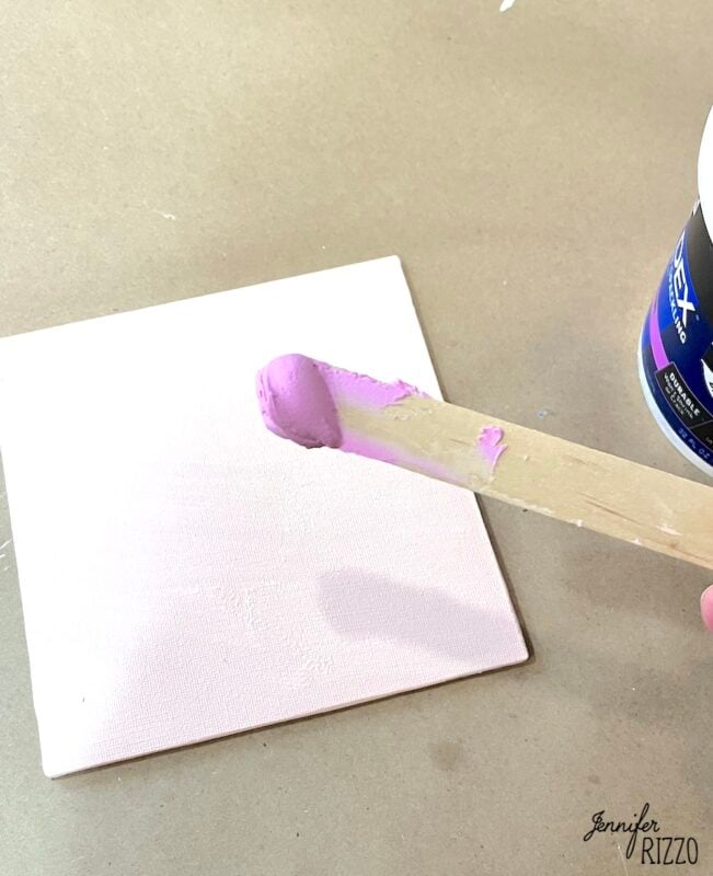 A hand holds a wooden stick with pink spackle over a small, square white canvas on a brown surface. A container of spackling paste is partially visible.