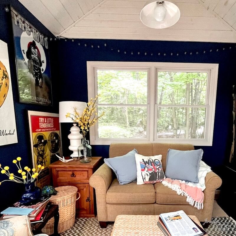 A cozy room with dark blue walls, a tan loveseat with pillows and a blanket, a window showing trees, posters on the wall, and a small side table with a vase of yellow flowers.
