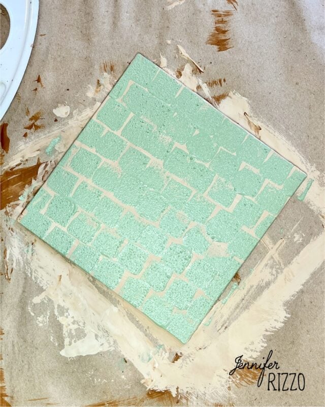 A square tile painted light green with a brick pattern sits on brown paper, surrounded by beige paint smudges; "Jennifer Rizzo" is written in the corner.