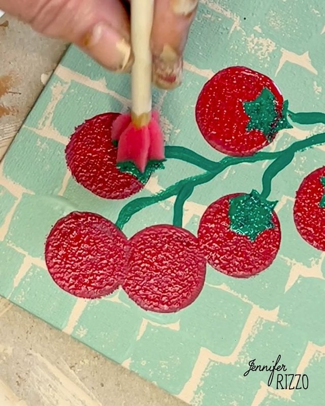 A hand uses a small paintbrush to add green paint to the stems of red tomatoes on a painted canvas with a light blue and white background.