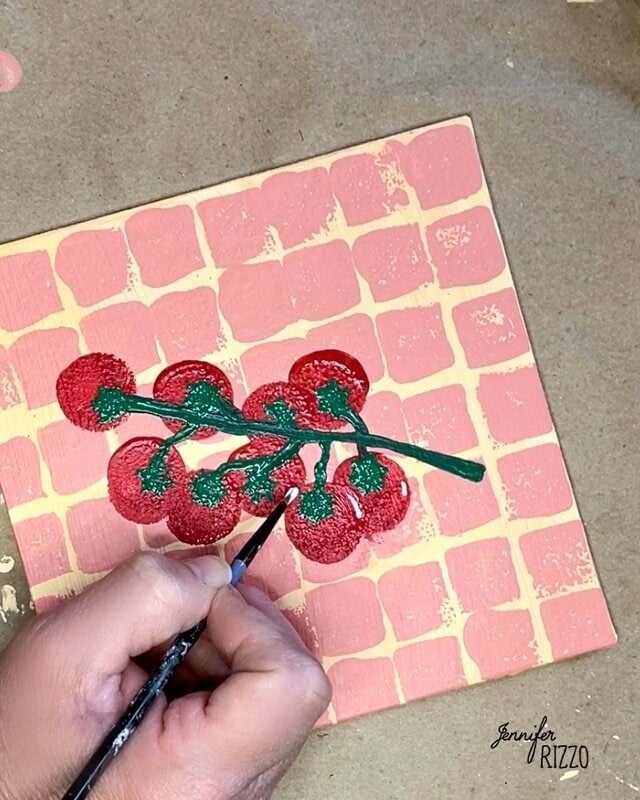 A person paints a branch of red berries with green leaves on a square tile featuring a pink and peach brick pattern. The artist's signature, Jennifer Rizzo, is visible.