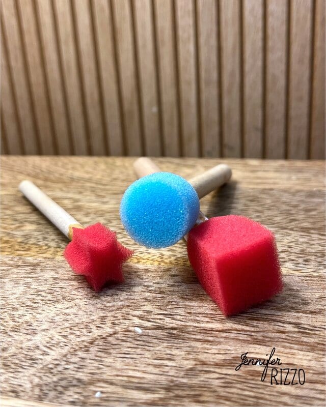 Three foam paint dabbers with wooden handles: a blue sphere, a red star, and a red cube, placed on a wooden surface with a ribbed background.