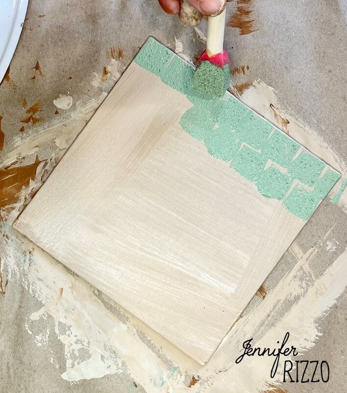 A hand uses a sponge brush to apply green paint to the top edge of a square wooden board on a paint-splattered surface.