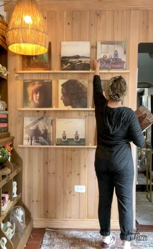 A person in dark clothing stands in front of wooden shelves displaying vinyl records, reaching up to adjust one of the records on the top shelf.