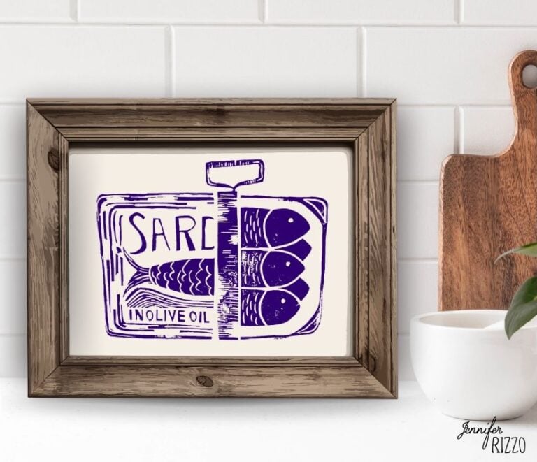 Framed artwork depicting a purple illustration of a sardine tin with three fish and the words "SARD IN OLIVE OIL," displayed on a kitchen counter.