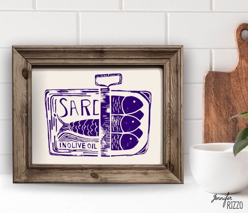 Framed artwork depicting a purple illustration of a sardine tin with three fish and the words "SARD IN OLIVE OIL," displayed on a kitchen counter.