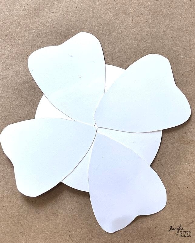 Four white, heart-shaped paper petals are arranged in a circular pattern on a brown background.