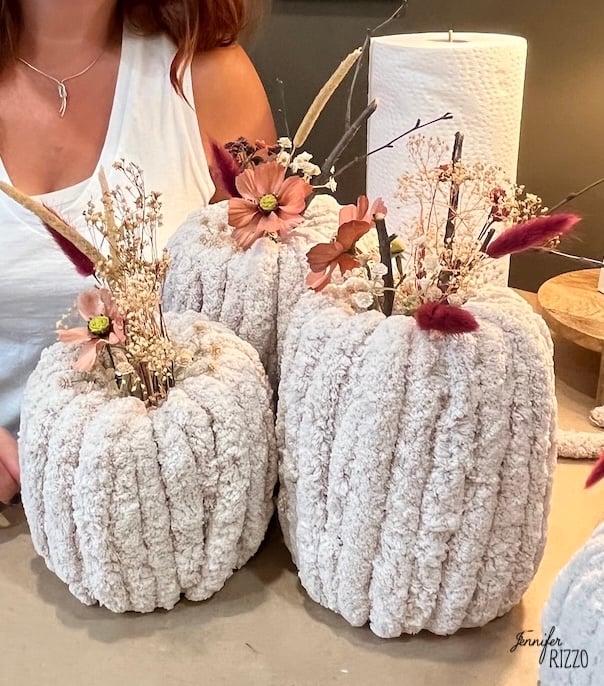 Three plush, textured fabric pumpkins decorated with dried flowers and foliage are displayed on a table. A person is partially visible in the background.