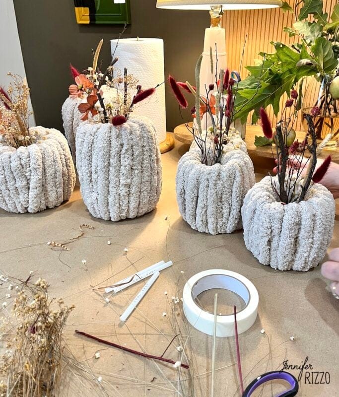 Several handmade textured vases filled with dried flowers are arranged on a table with crafting supplies, including tape, scissors, sticks, and loose dried botanicals.