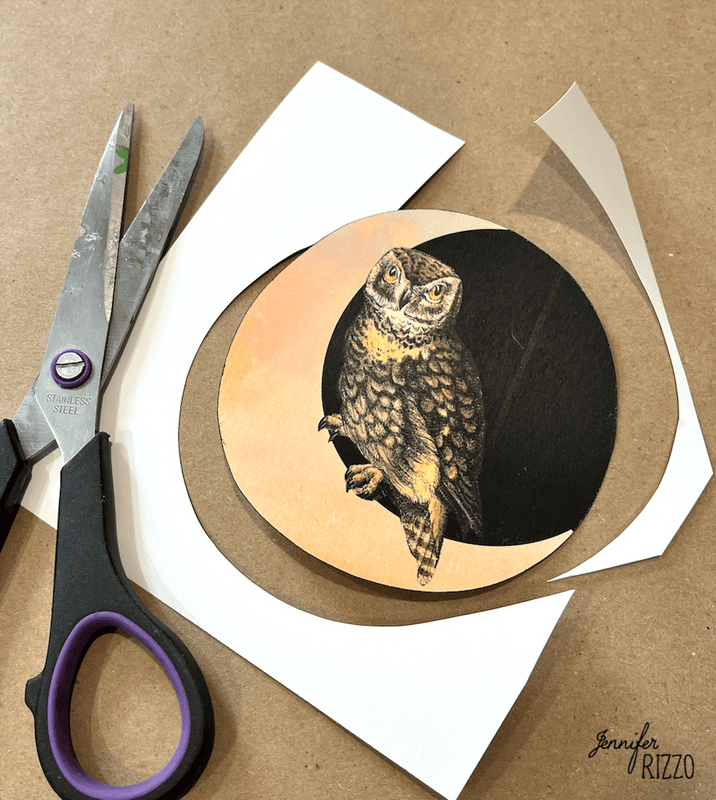 A pair of scissors and a circular cutout featuring an owl illustration are on a brown surface, with scraps of white paper nearby.