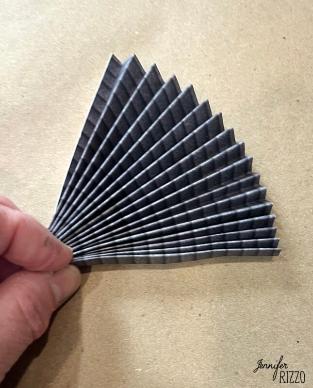 A hand holding a piece of black paper folded in an accordion style, forming a fan shape, on a brown background.