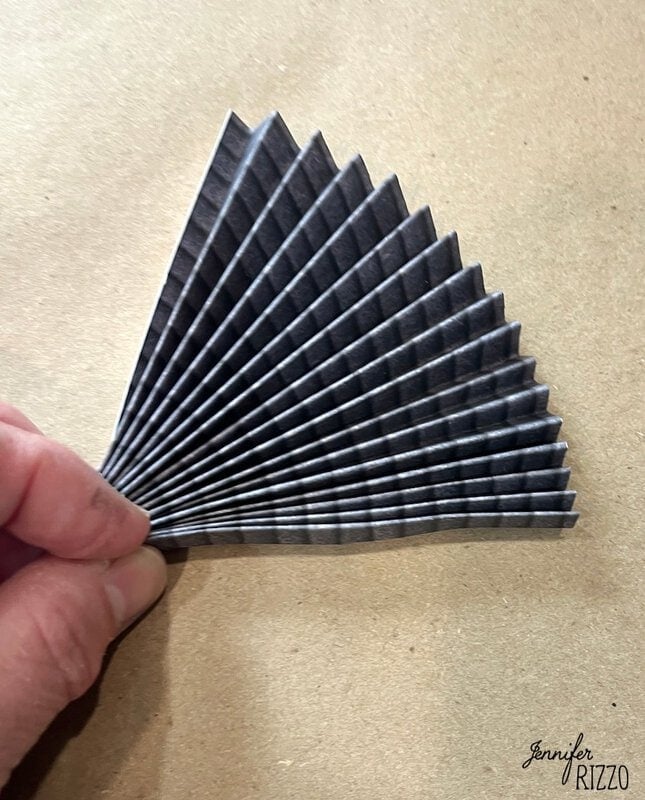 A hand holding a piece of black paper folded in an accordion style, forming a fan shape, on a brown background.