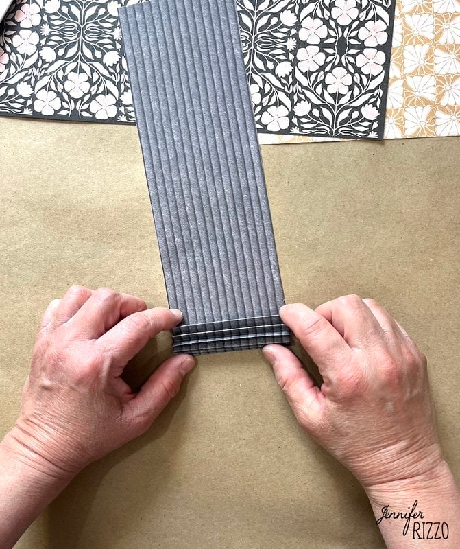 Two hands folding a piece of gray, corrugated paper on a brown surface, with patterned paper sheets in the background.