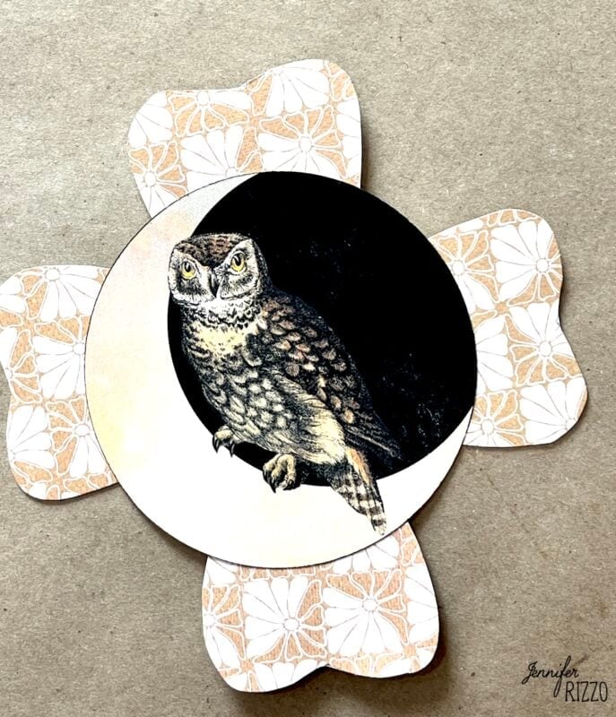 A circular card featuring an illustrated owl is placed on top of a flower-shaped, patterned paper background.