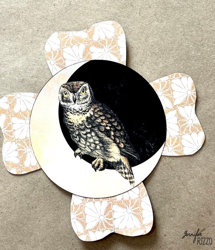 A circular card featuring an illustrated owl is placed on top of a flower-shaped, patterned paper background.
